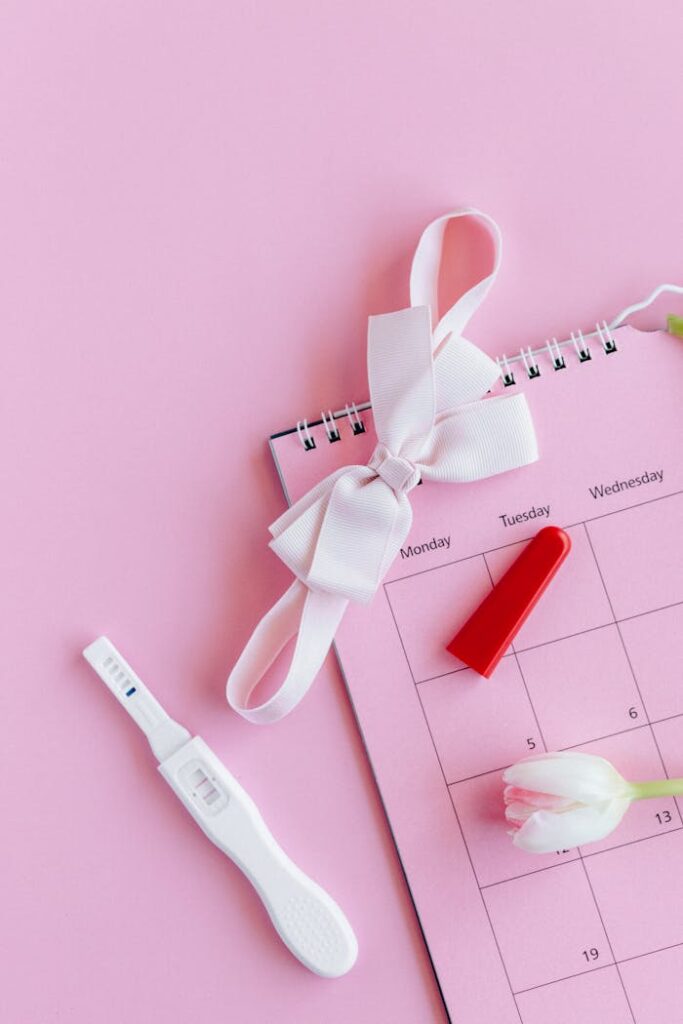 Flat lay of a pregnancy test on a pink background with a calendar and bow.